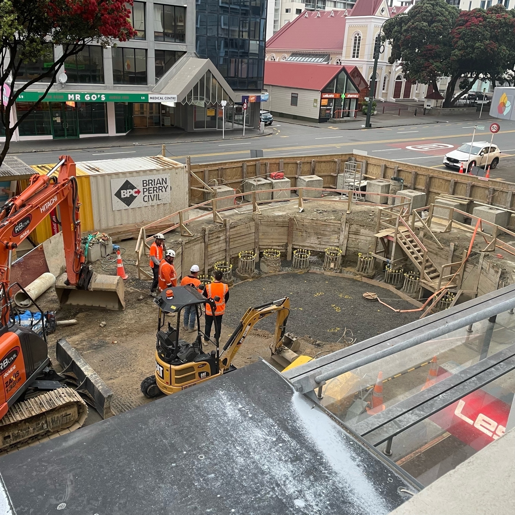 Four men in hi-visibility jackets inspect a large reinforced hole in the ground. They are surrounded by digging machinery and safety fences. Taranaki St, Wellington and the Methodist Church in the background. 