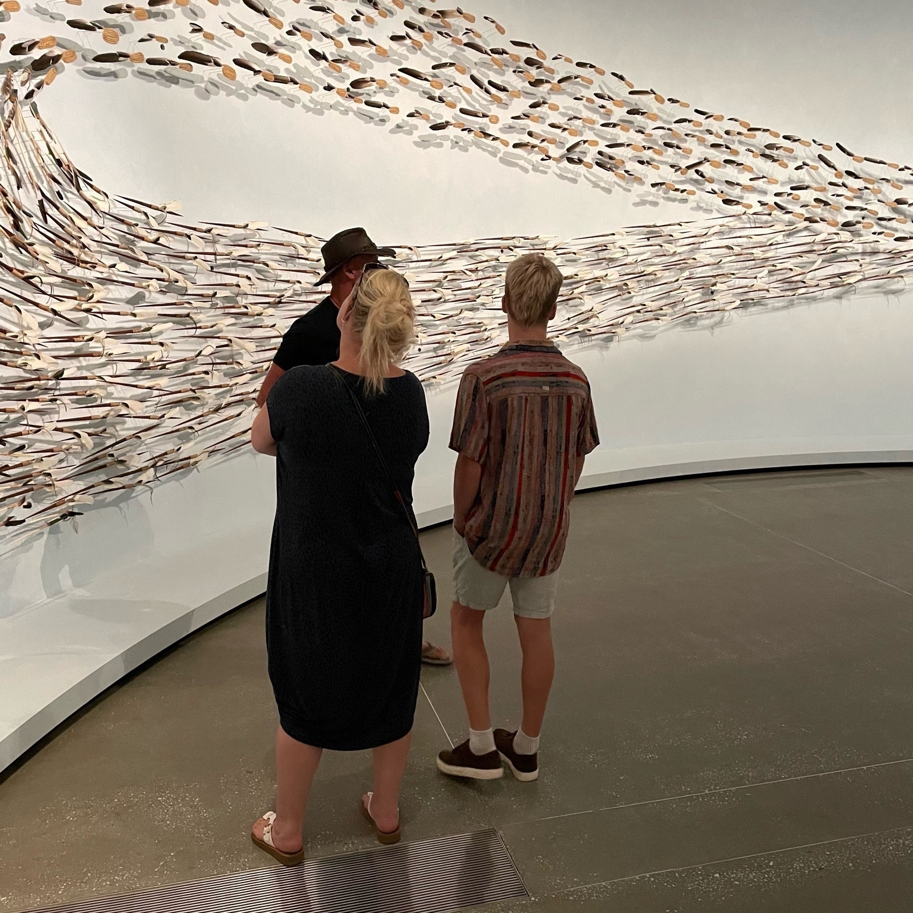 Three people looking at an installation of thousands of feathers, shells and other natural objects that form an ellipsis reminiscent of a murmuration of birds