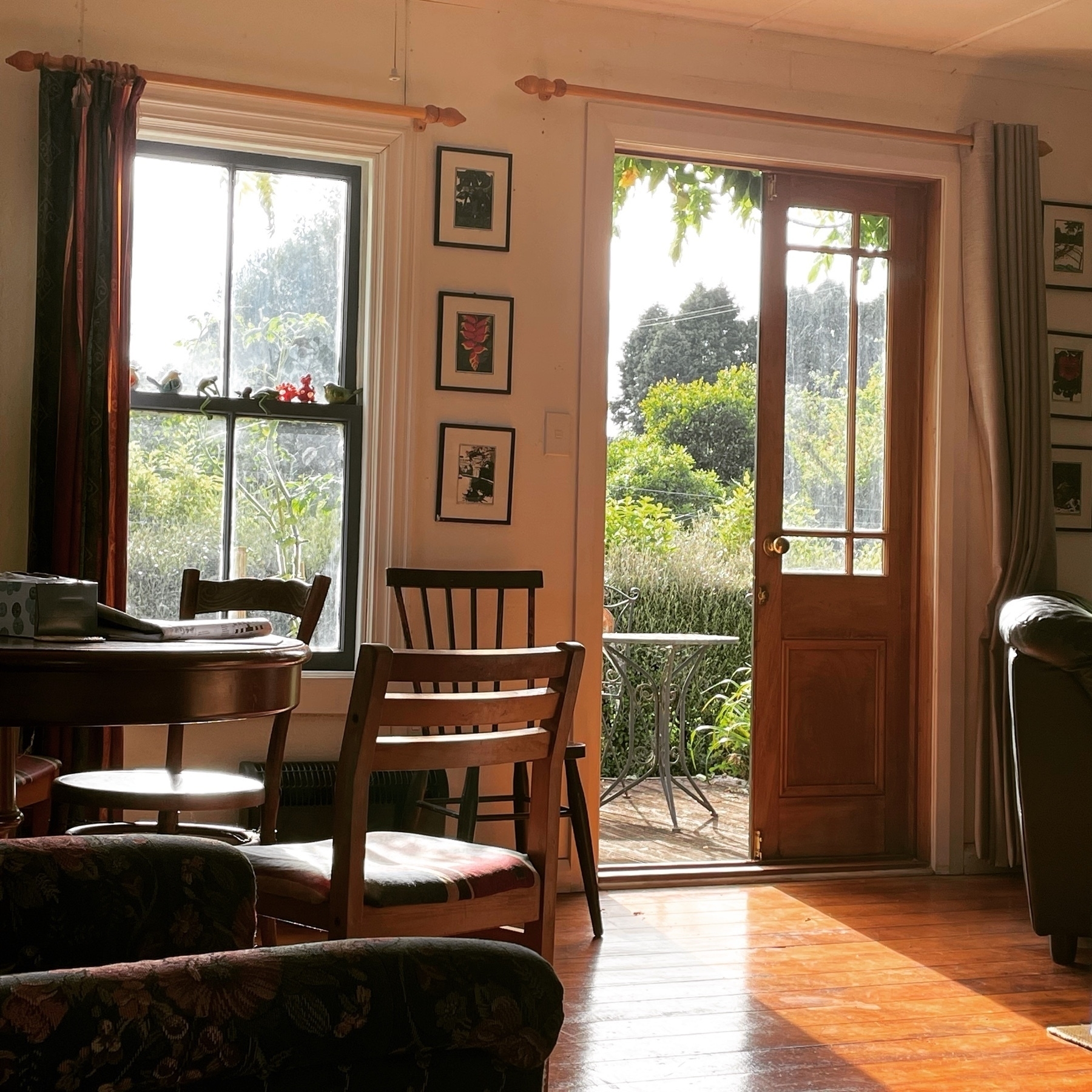 Morning light streams through an open French door and window in a wooden country cottage onto chairs and a table. A garden is visible outside. 